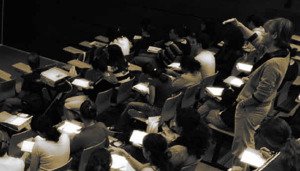 Vista de una persona docente de pie, en medio de una clase, en actitud de impartir clase.