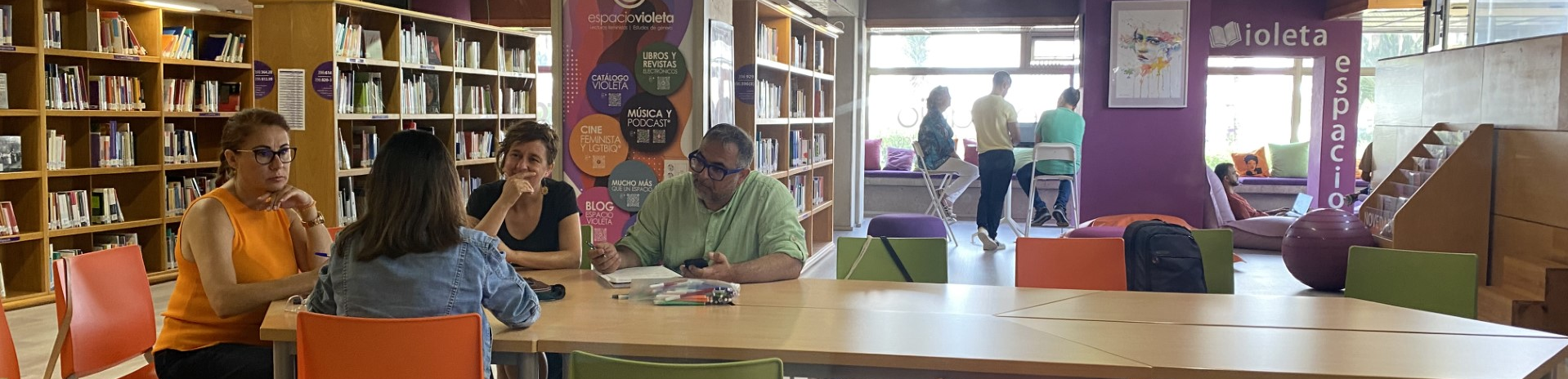 Un grupo de 4 personas trabaja en torno a una mesa en una biblioteca. Al fondo, otras 3 personas trabajan en torno a otra mesa alta. Otra persona trabaja en un ordenador portátil sentado en cojines en el suelo.