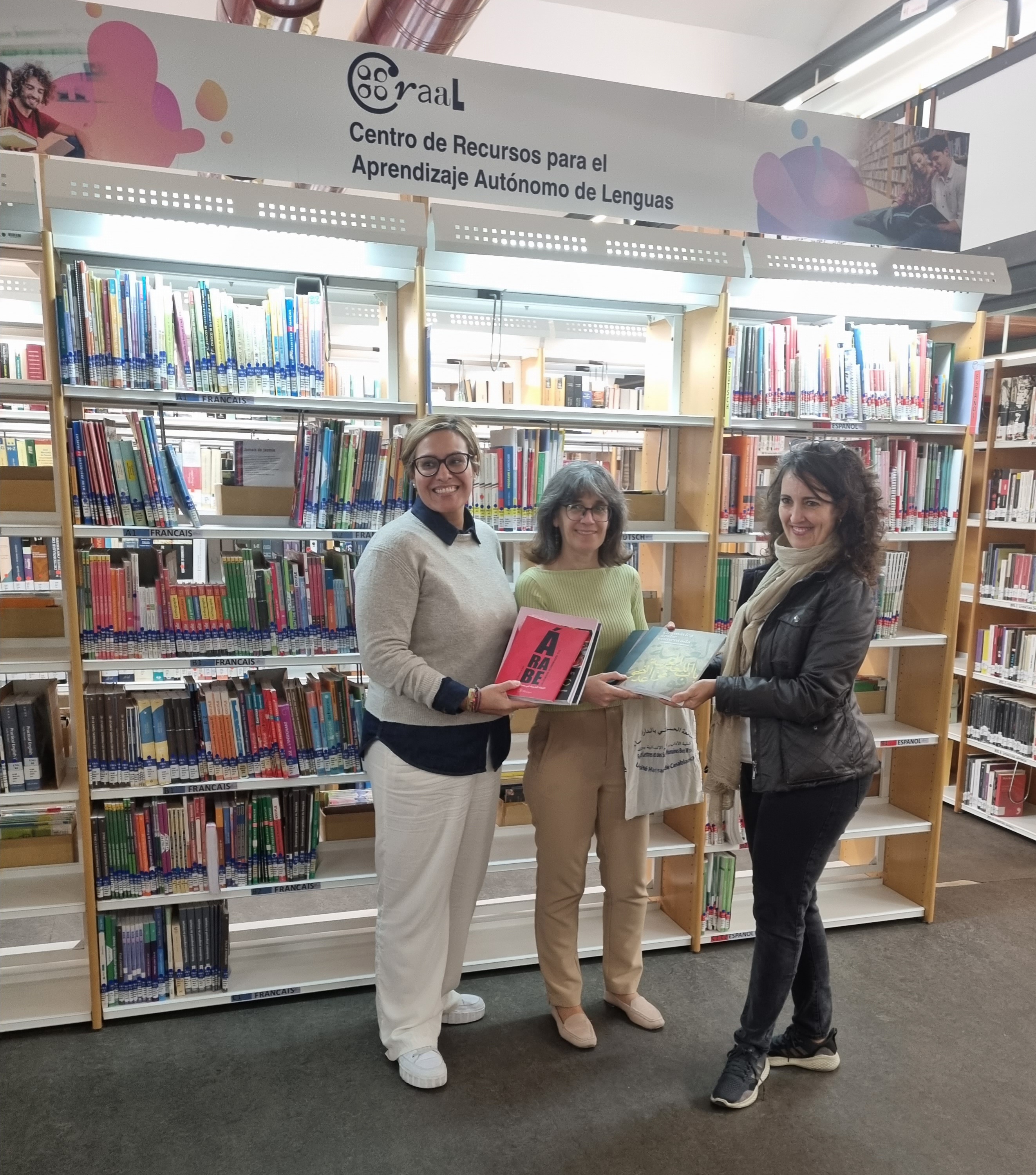 Foto de grupo de tres mujeres mostrando libros frente a estantes de biblioteca.De izquierda a derecha: Cristina Alfonzo de Tovar, coordinadora del CRAAL, Ana Ruth Vidal Luengo, responsable del área de árabe del CraaL, y Teresa Cabrera Morales, de la Biblioteca Universitaria