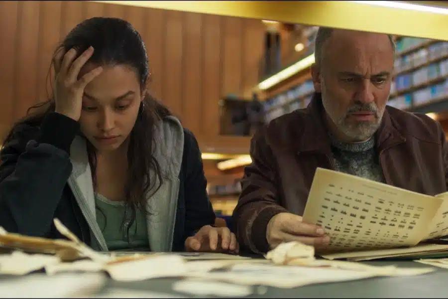 Dos personas, en primer plano, estudiando en una biblioteca. Al fondo, otras personas lectoras y estanterías.