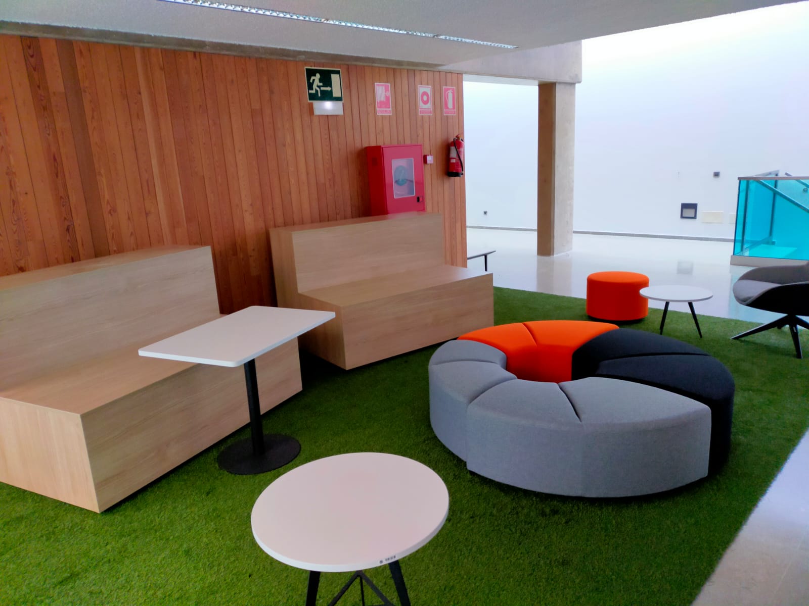 Vista de zona de césped artificial de la biblioteca, con pufs, sillón, mesa y 2 mesitas y dos gradas de madera.