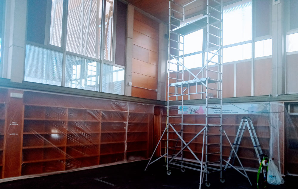Andamio y material de obra en sala de biblioteca vacía, con polvo en el ambiente.